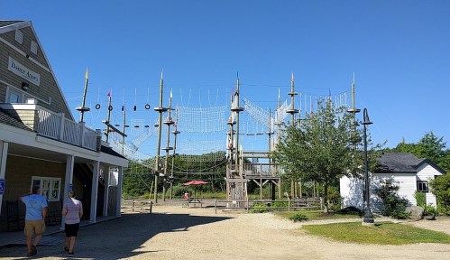 A ropes course with towers and obstacles, set against a clear blue sky, near a building and walking path.