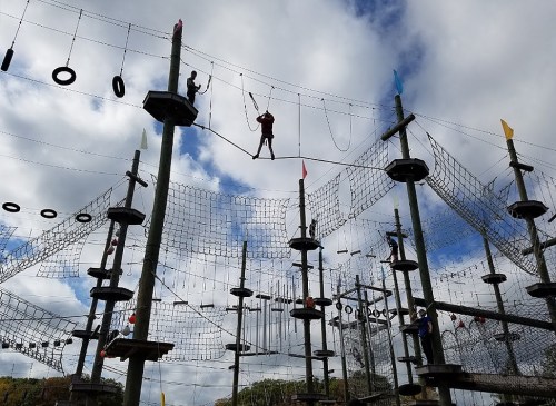 Aerial obstacle course with people navigating ropes and nets against a cloudy sky.