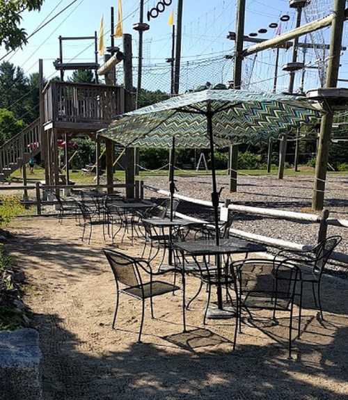 Outdoor seating area with tables and chairs under a patterned umbrella, near a ropes course and trees.