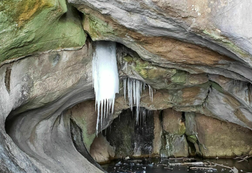 A cave interior with icicles hanging from the ceiling and moss-covered rocks, reflecting in still water below.