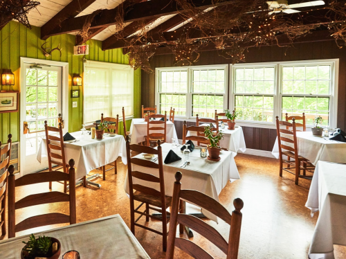 Cozy restaurant interior with wooden chairs, tables set with white tablecloths, and large windows letting in natural light.