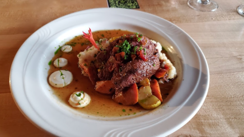 A plate of tender meat atop mashed potatoes, surrounded by colorful vegetables and garnished with herbs.
