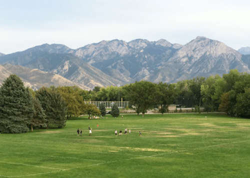 A grassy field with people playing in the foreground, surrounded by trees and mountains in the background.