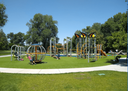 A vibrant playground with various equipment, including swings and climbing structures, set in a green park under a clear blue sky.
