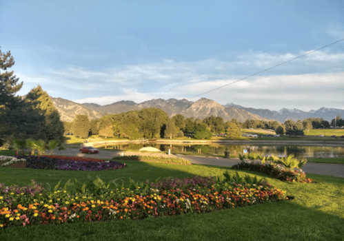 Scenic view of a park with colorful flowers, a calm lake, and mountains in the background under a clear sky.