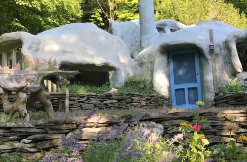 A whimsical, white, cave-like structure surrounded by greenery and colorful flowers, featuring a blue door and stone pathways.