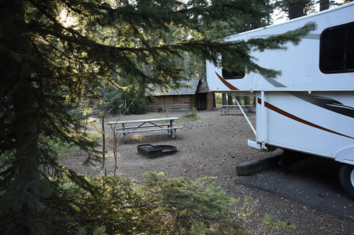 A camper parked near a picnic area surrounded by trees, with a fire pit and cabin in the background.