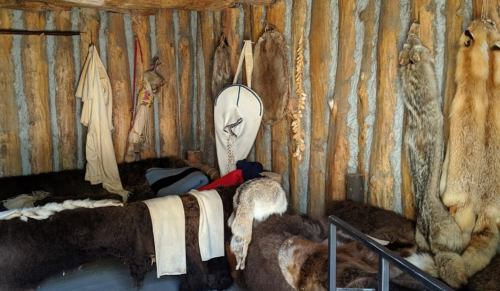 Interior of a rustic cabin with fur-covered bedding, animal pelts on the walls, and traditional clothing hanging.