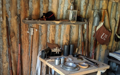 A rustic wooden shelf and table display various historical tools, utensils, and weapons against a log wall backdrop.