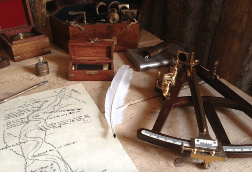 A wooden table with vintage navigation tools, a feather quill, and a map featuring a winding river.