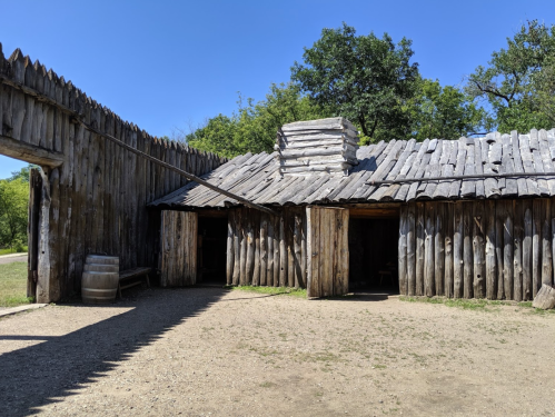 A rustic wooden structure with a slanted roof, surrounded by a wooden fence and trees under a clear blue sky.