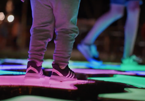 A child's feet in gray sweatpants and black sneakers stand on colorful, illuminated tiles at night.