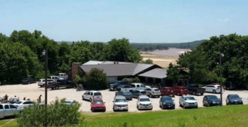 A parking lot filled with vehicles near a building, surrounded by trees and a river in the background on a sunny day.