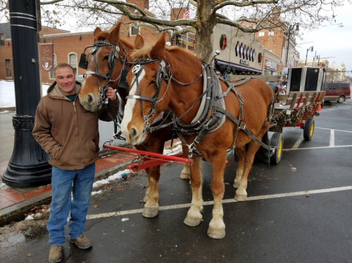 A man stands beside two brown horses hitched to a cart in a snowy town square.