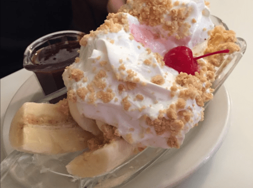 A banana split topped with whipped cream, crushed cookies, a cherry, and a side of chocolate sauce.