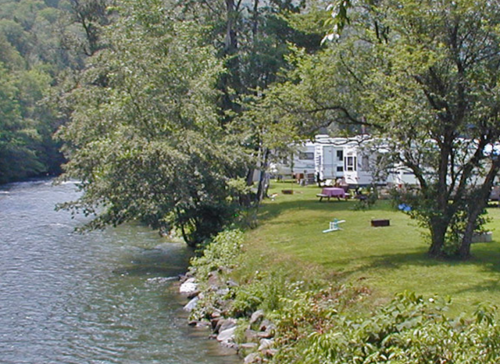 A serene riverside scene featuring a grassy area with a trailer, picnic table, and trees along the water's edge.