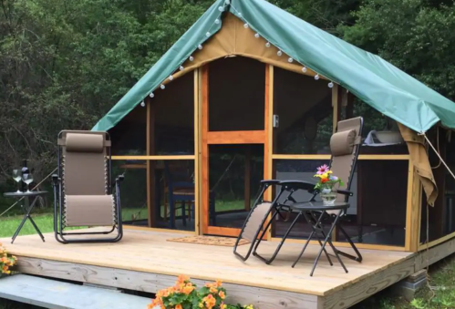 A cozy glamping tent on a wooden deck, with two chairs, a small table, and flowers, surrounded by greenery.
