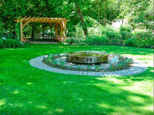 A circular stone garden with a water feature, surrounded by flowers and a wooden pergola in a lush green setting.