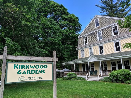 Sign for Kirkwood Gardens at Squam Lakes Natural Science Center, with a large house and green trees in the background.