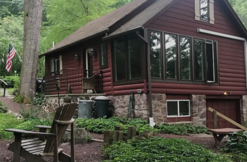 A red log cabin surrounded by greenery, featuring large windows and an American flag. Outdoor seating is visible in front.