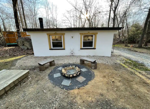 A small white building with two windows, surrounded by trees and a fire pit with stone seating in front.