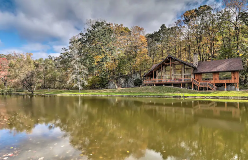 A rustic cabin by a serene pond, surrounded by colorful autumn trees under a partly cloudy sky.