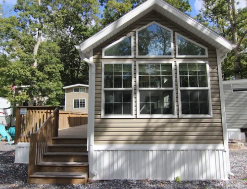 A modern cabin with large windows and a wooden deck, surrounded by trees and gravel.