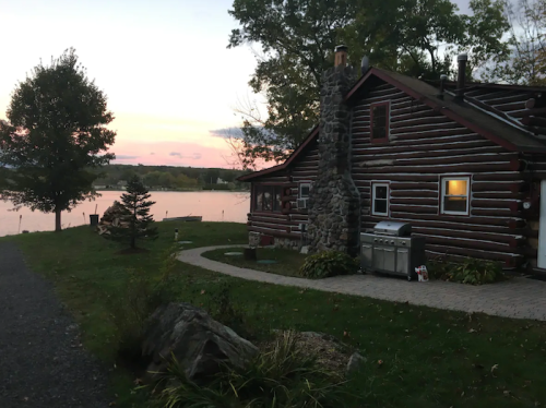 A rustic log cabin by a lake at sunset, with a stone chimney and grill, surrounded by trees and a grassy area.