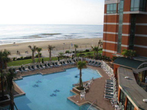 A beachfront hotel pool with lounge chairs, overlooking the ocean and sandy beach under a clear sky.