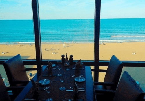 A dining table set for a meal, overlooking a beach and ocean through large windows.