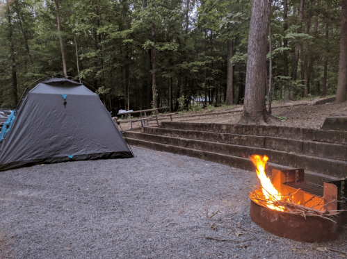 A camping scene with a tent, a fire pit with flames, and a wooded area in the background.