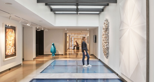 A modern art gallery featuring a woman walking on glass floors, surrounded by various artworks and sculptures.