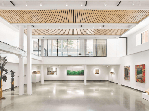 A spacious art gallery with high ceilings, wooden beams, and various artworks displayed on white walls.