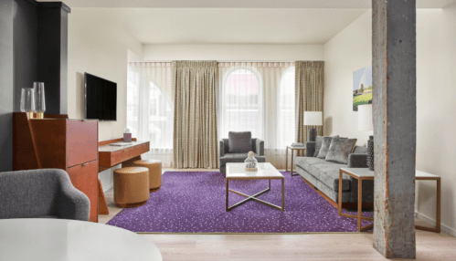 Modern living room with a gray sofa, armchairs, a coffee table, and large windows, featuring purple carpet and stylish decor.