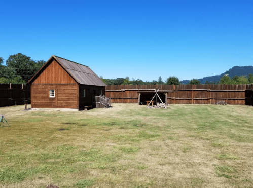 A wooden building surrounded by a tall wooden fence, with a grassy area and mountains in the background under a clear blue sky.