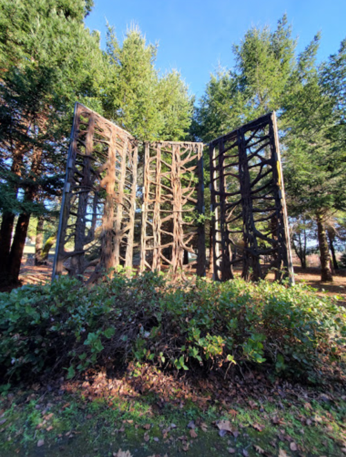 A large, abstract wooden sculpture resembling tree roots, surrounded by greenery and tall trees under a clear blue sky.
