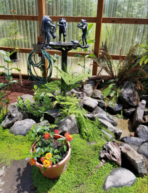A garden scene featuring a sculpture of three figures, surrounded by rocks, ferns, and a potted flower plant.