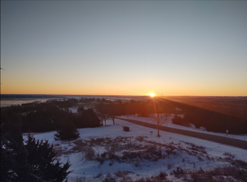 A serene winter landscape at sunset, with snow-covered ground and trees silhouetted against a colorful sky.