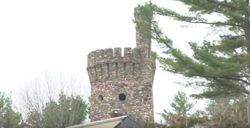 A stone tower with battlements rises above trees, set against a cloudy sky.