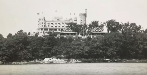 Black and white image of a large castle-like building on a rocky shore, surrounded by trees and water.