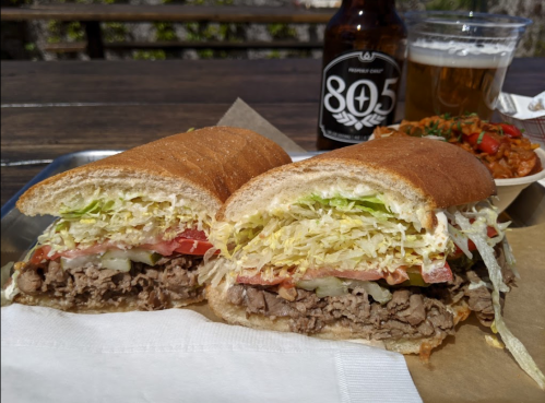 A close-up of a sandwich filled with beef, lettuce, and tomato, served with a beer and a side of beans.