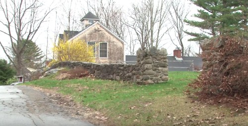 A rustic house with a stone wall and trees, set in a rural landscape on a cloudy day.