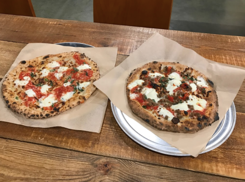 Two pizzas on metal trays, one topped with red sauce and mozzarella, the other with red sauce, mozzarella, and herbs.