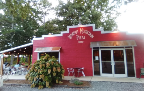 Red brick building with "Lookout Mountain Pizza" sign, outdoor seating, and trees in the background. Evening ambiance.