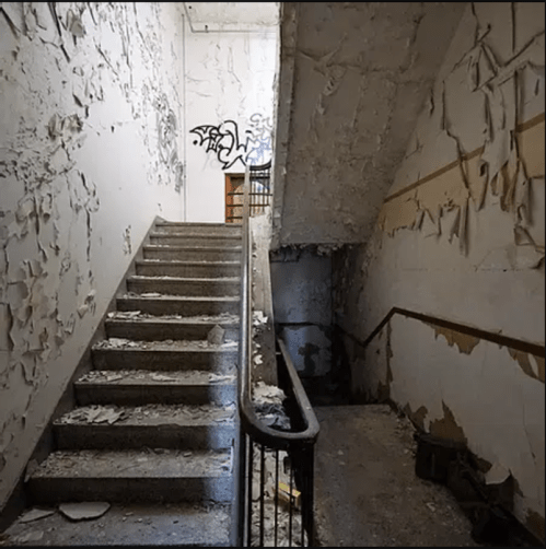 A worn staircase in a dilapidated building, with peeling paint and graffiti on the walls.