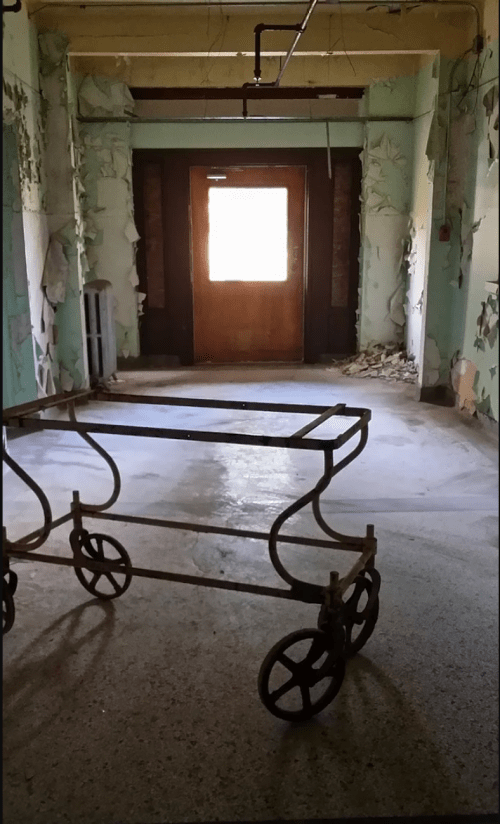 An abandoned hallway with peeling wallpaper and a vintage cart in the foreground, leading to a door with light.