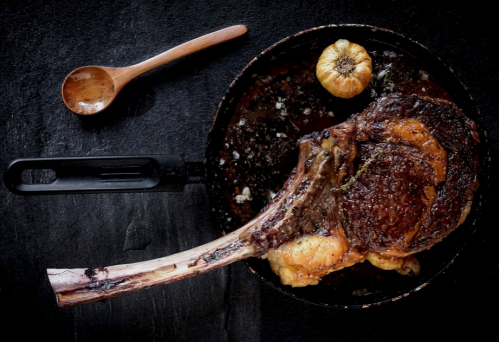 A large, cooked tomahawk steak in a skillet, garnished with a roasted garlic bulb and a wooden spoon nearby.