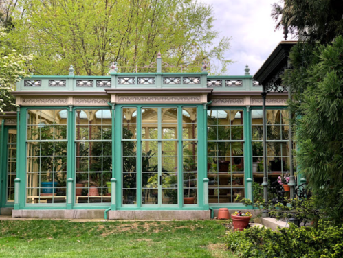 A green glass conservatory surrounded by greenery, featuring large windows and potted plants.