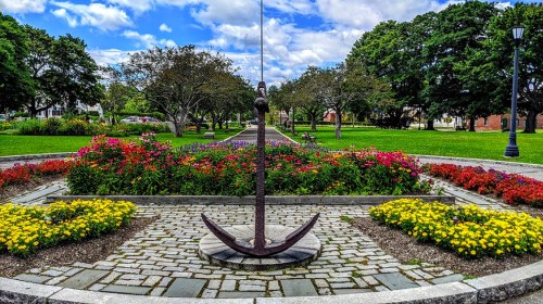 A park scene featuring a large anchor sculpture surrounded by colorful flower beds and green trees under a blue sky.