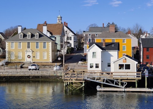 Colorful buildings line a waterfront, reflecting in the calm water under a clear blue sky.
